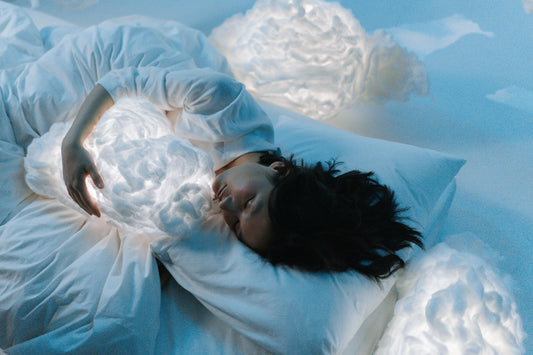 A person sleeps peacefully in bed, holding a glowing, cloud-shaped pillow, surrounded by other softly lit clouds, creating a dreamy, serene atmosphere